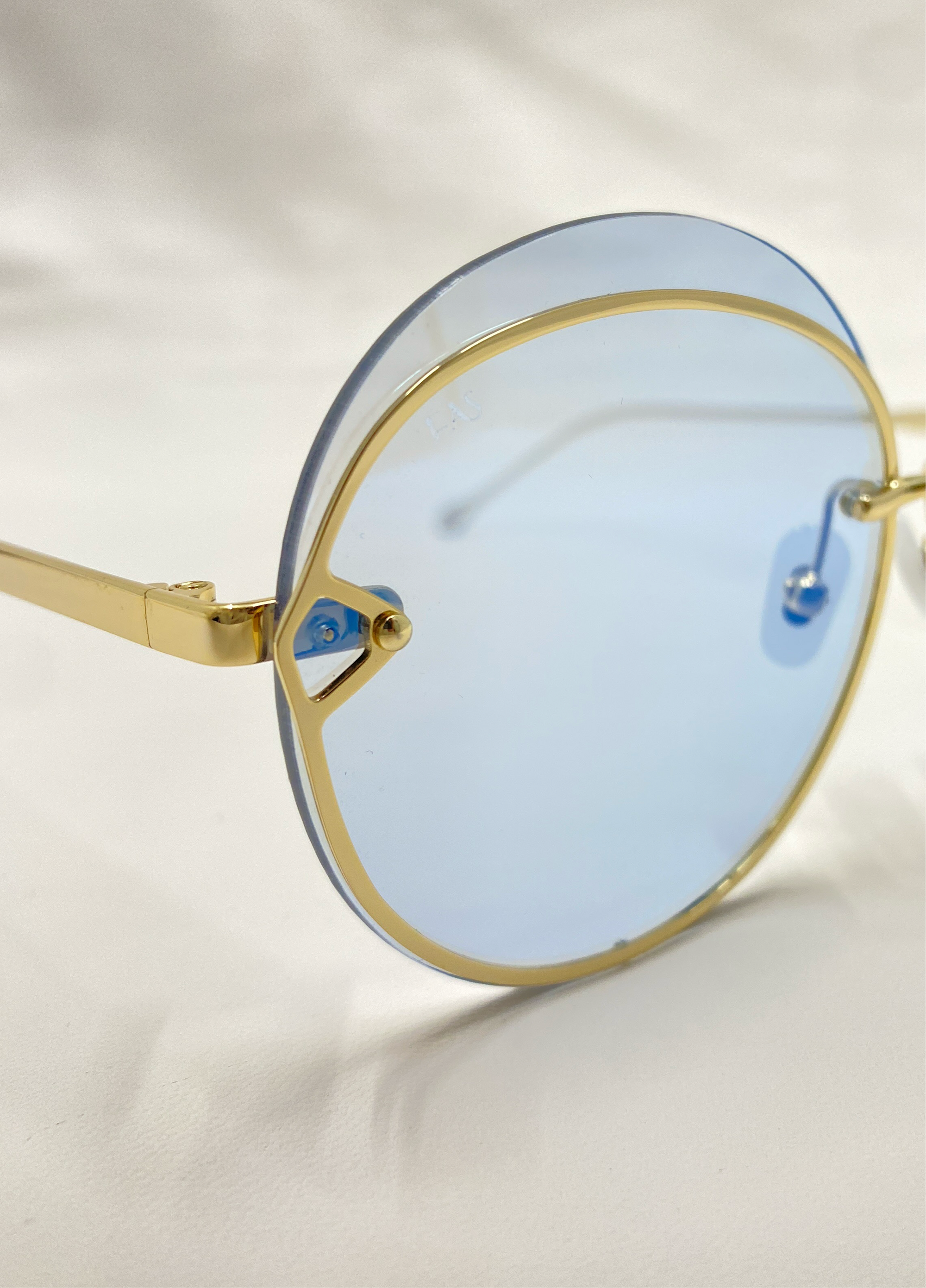 Close-up of a pair of For Art&#39;s Sake® Quartz round sunglasses with 18-karat gold-plated frames and light blue-tinted Nylon lenses, resting on a white surface. The nose pads and hinges are clearly visible, highlighting the sleek design and detailing of the eyewear.