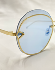 Close-up of a pair of For Art's Sake® Quartz round sunglasses with 18-karat gold-plated frames and light blue-tinted Nylon lenses, resting on a white surface. The nose pads and hinges are clearly visible, highlighting the sleek design and detailing of the eyewear.