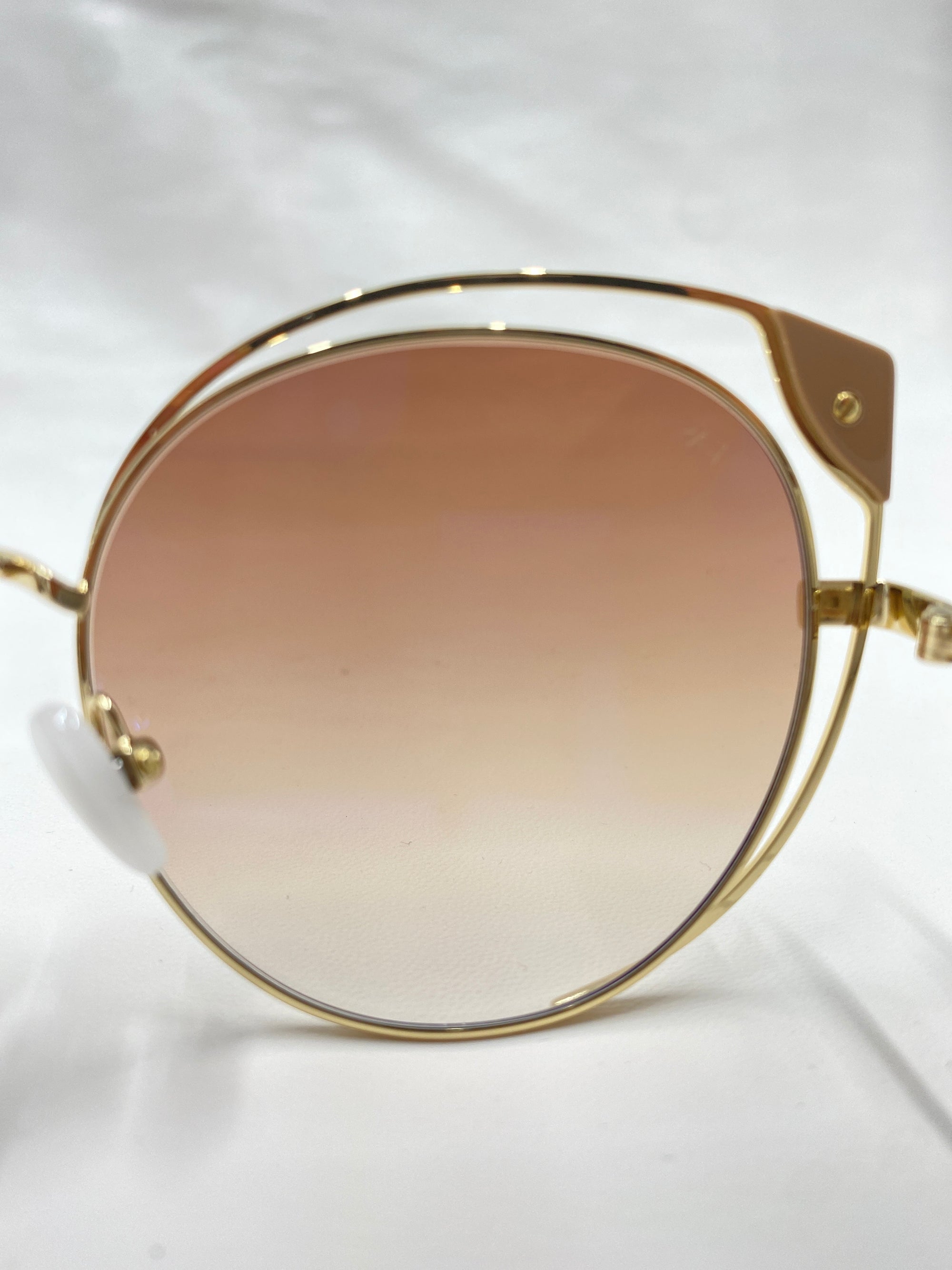 Close-up view of a stylish pair of Skylar cat-eye silhouette sunglasses by For Art's Sake® with a gold frame and gradient nylon lenses. The UVA UVB-protected glasses are set against an out-of-focus light surface, highlighting their elegant design and details.