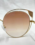 Close-up view of a stylish pair of Skylar cat-eye silhouette sunglasses by For Art's Sake® with a gold frame and gradient nylon lenses. The UVA UVB-protected glasses are set against an out-of-focus light surface, highlighting their elegant design and details.