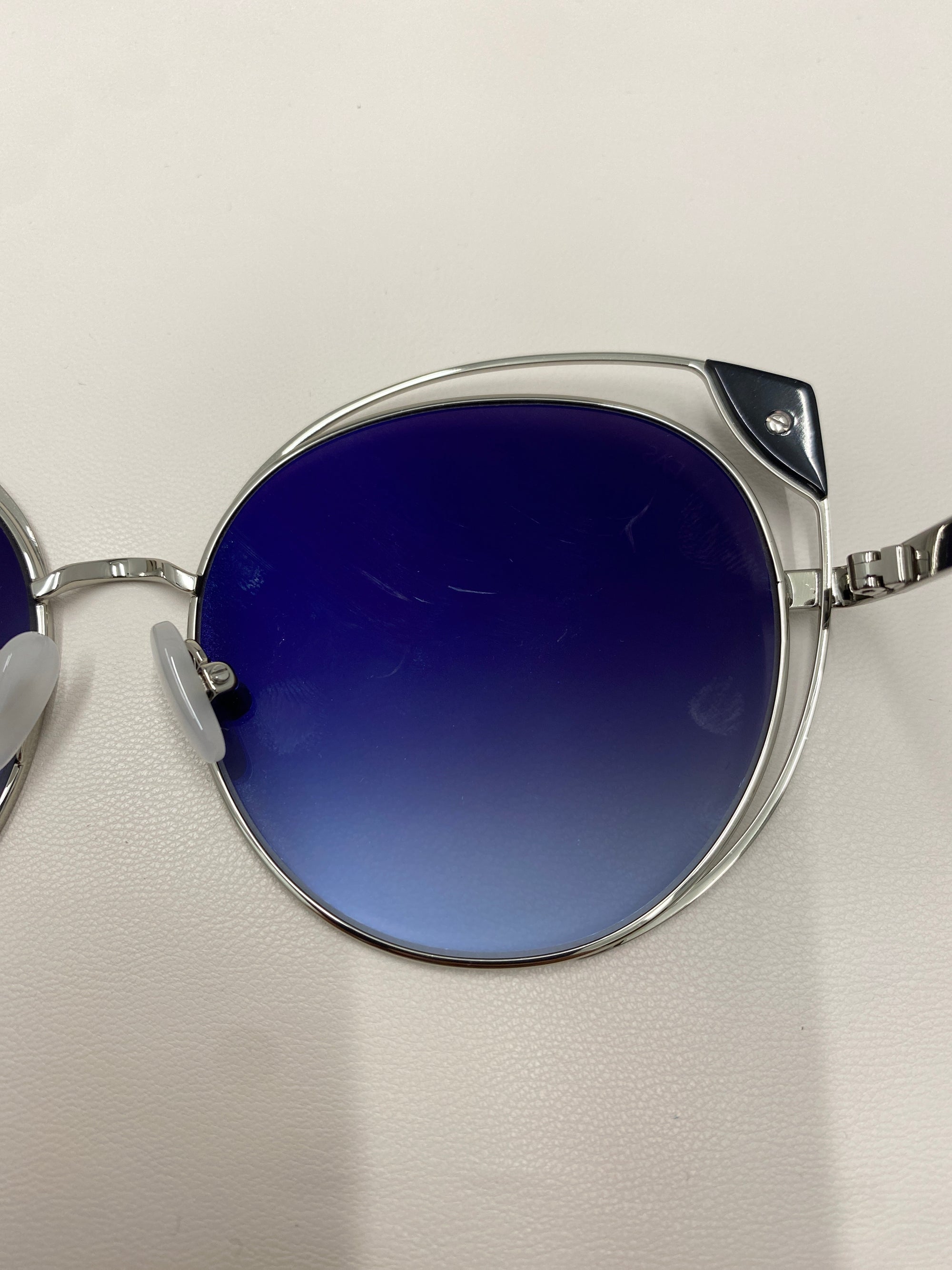 Close-up of Skylar, a pair of stylish sunglasses by For Art's Sake®, featuring silver metal frames and large, round lenses with a blue gradient tint. The UVA UVB-protected sunglasses are resting on a white surface, showcasing the hinge connecting the lens to the arm on the right side of the image.