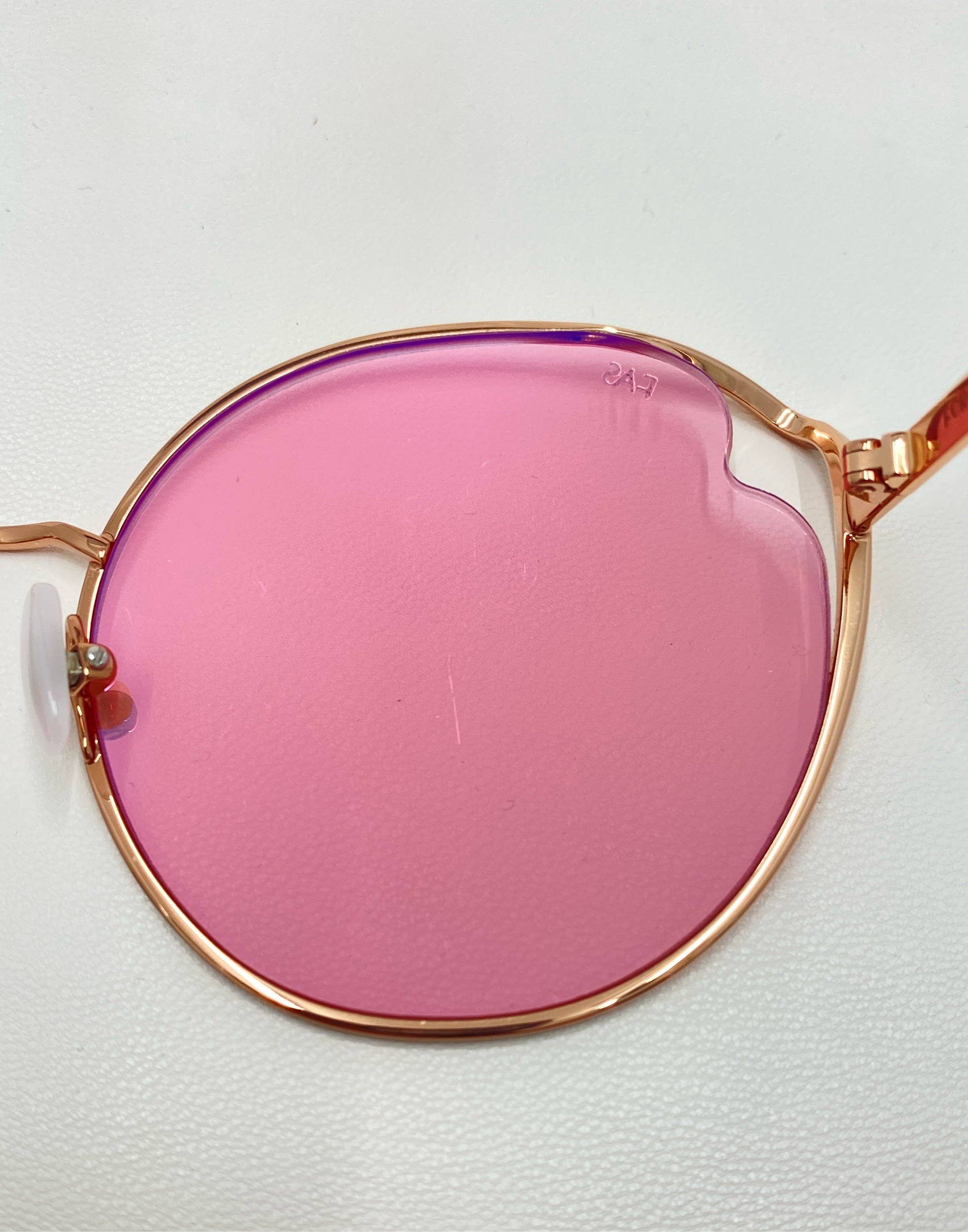 Close-up of a rose gold metal frame eyeglass named &quot;Cat&quot; by For Art&#39;s Sake®, featuring a pink nylon lens with UVA &amp; UVB protection. The lens displays a small &quot;247&quot; imprint on its surface. The eyeglass, which includes adjustable nosepads, rests on a white background.