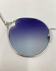 Close-up image of the Cat sunglasses by For Art's Sake®, featuring stylish thin silver frames and gradient blue to white, UVA & UVB-protected nylon lenses, placed on a white surface. The photo highlights the elegant design, color transition of the lenses, and adjustable nosepads for added comfort.