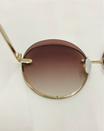 A close-up image of the Quartz sunglasses by For Art's Sake®, featuring 18-karat gold-plated frames and gradient lenses that transition from dark brown at the top to a lighter shade at the bottom. The Quartz sunglasses are lying on a white surface.