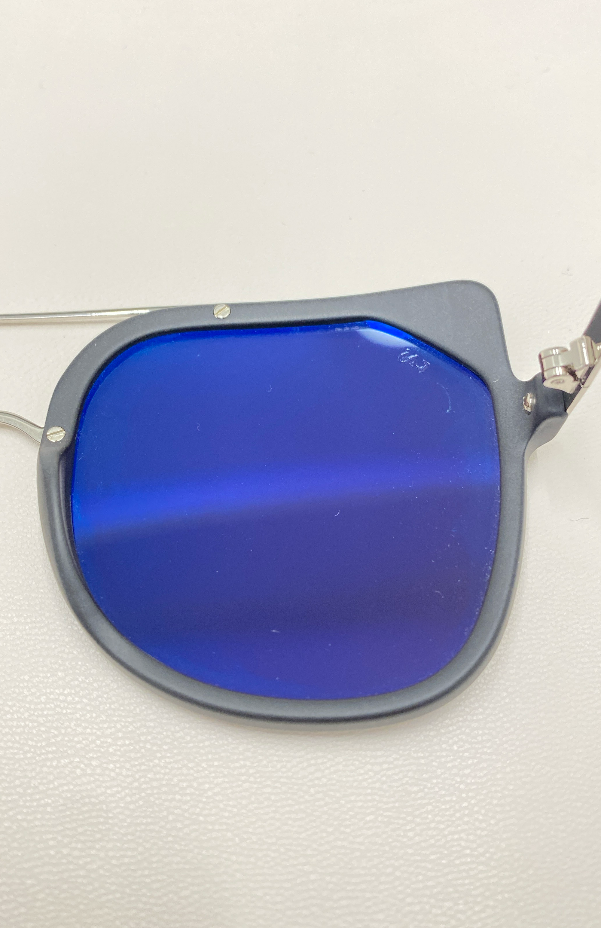 Close-up image of the Big Game lens from For Art&#39;s Sake® sunglasses. The rectangular lens with rounded corners is tinted a deep, reflective blue and offers UV protection. Partially attached to a silver frame, the rest of the frame is not visible. The background is plain white.