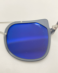 Close-up image of the Big Game lens from For Art's Sake® sunglasses. The rectangular lens with rounded corners is tinted a deep, reflective blue and offers UV protection. Partially attached to a silver frame, the rest of the frame is not visible. The background is plain white.