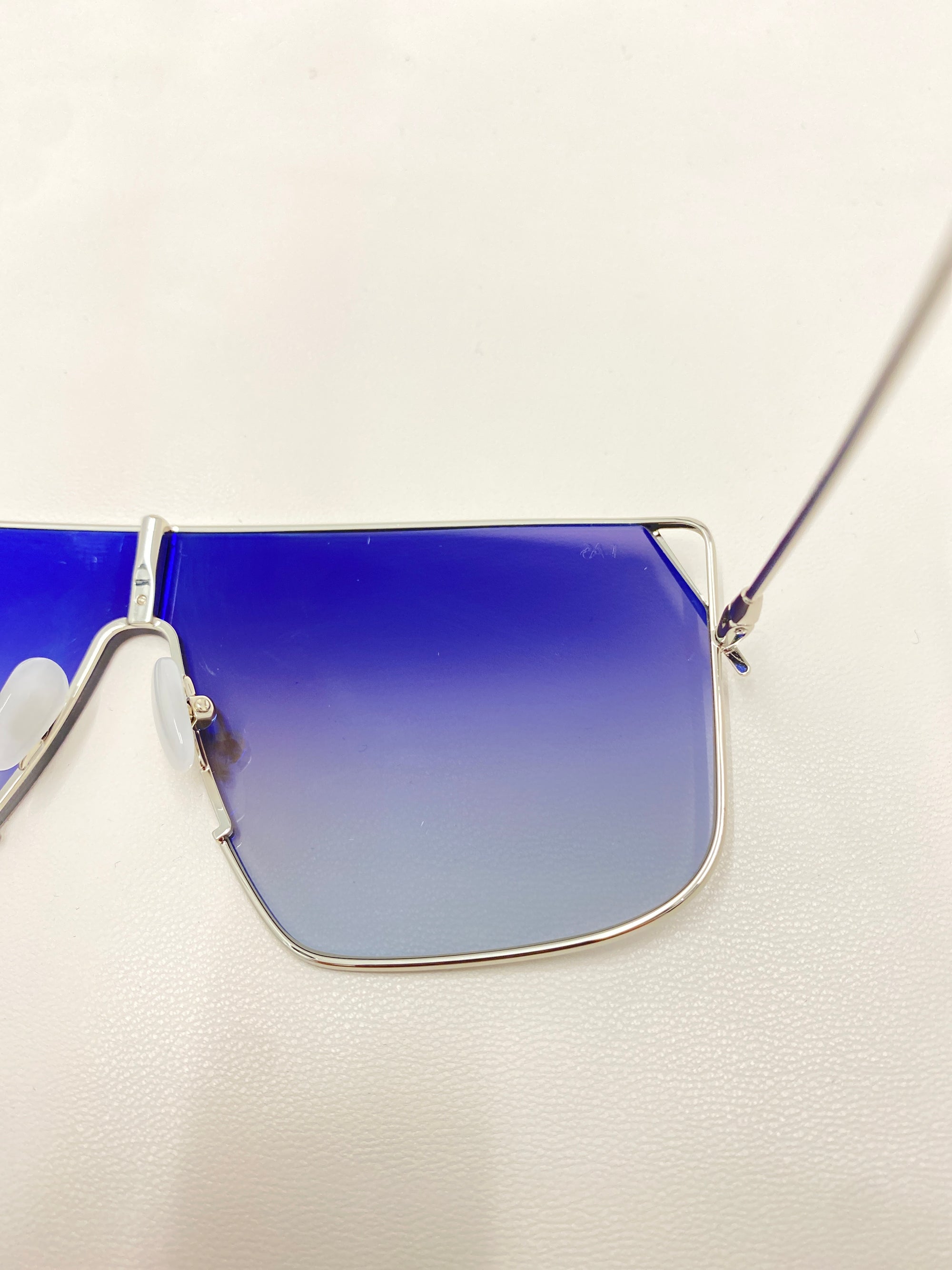 Close-up of the For Art&#39;s Sake® Horizon sunglasses featuring blue gradient nylon lenses and a thin metallic frame. The left lens is fully visible, while the right lens is partly shown. These stylish shades offer complete UVA &amp; UVB protection against harmful rays, all set against a plain, light surface backdrop.