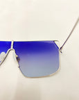 Close-up of the For Art's Sake® Horizon sunglasses featuring blue gradient nylon lenses and a thin metallic frame. The left lens is fully visible, while the right lens is partly shown. These stylish shades offer complete UVA & UVB protection against harmful rays, all set against a plain, light surface backdrop.