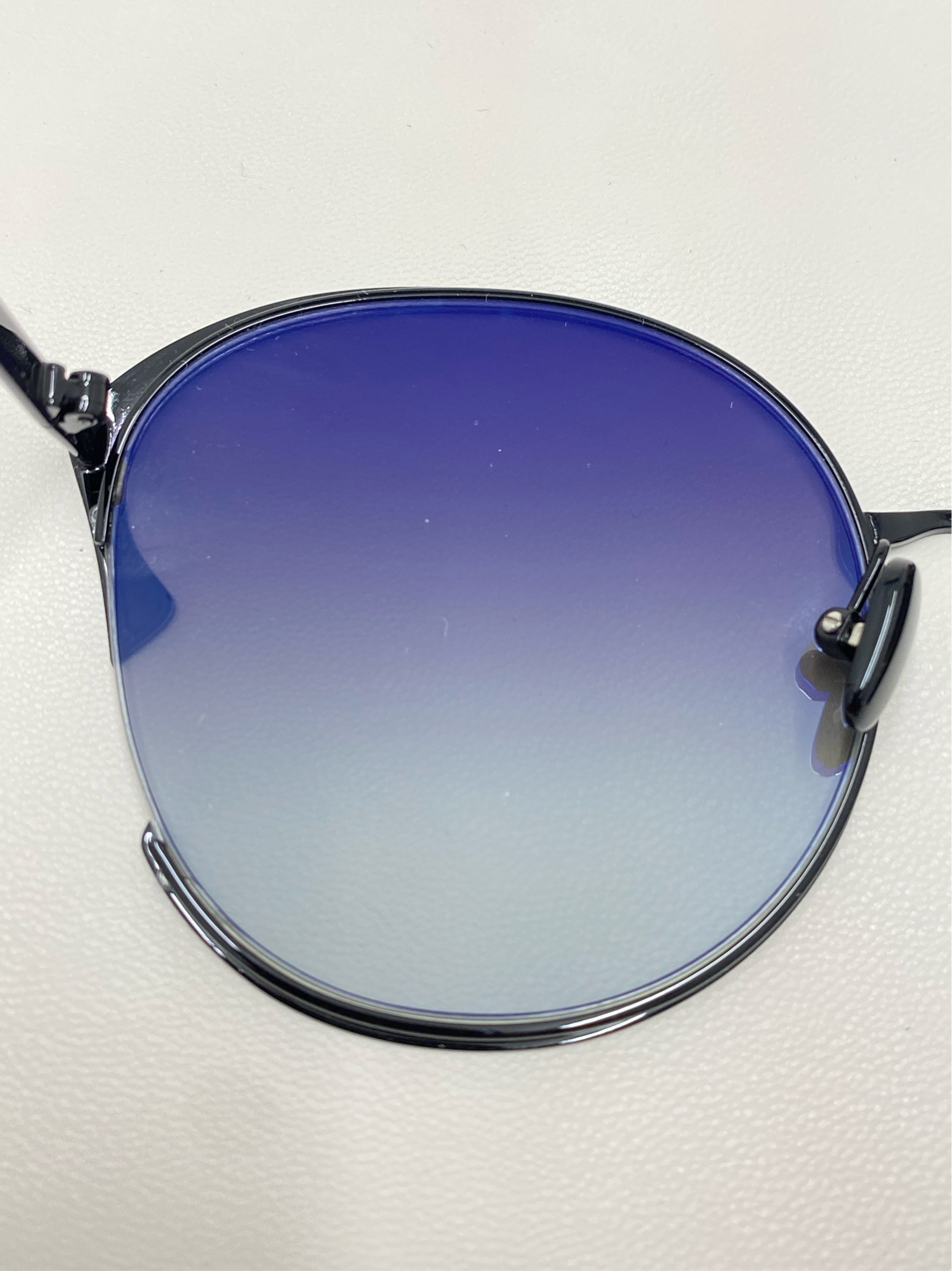 Close-up shot of a single lens of the Alectrona sunglasses by For Art's Sake®, featuring a gradient tint that transitions from dark blue at the top to a lighter color at the bottom. The metal frame is visible on the sides. These sunglasses boast UVA & UVB-protected nylon lenses, all set against a plain white background.