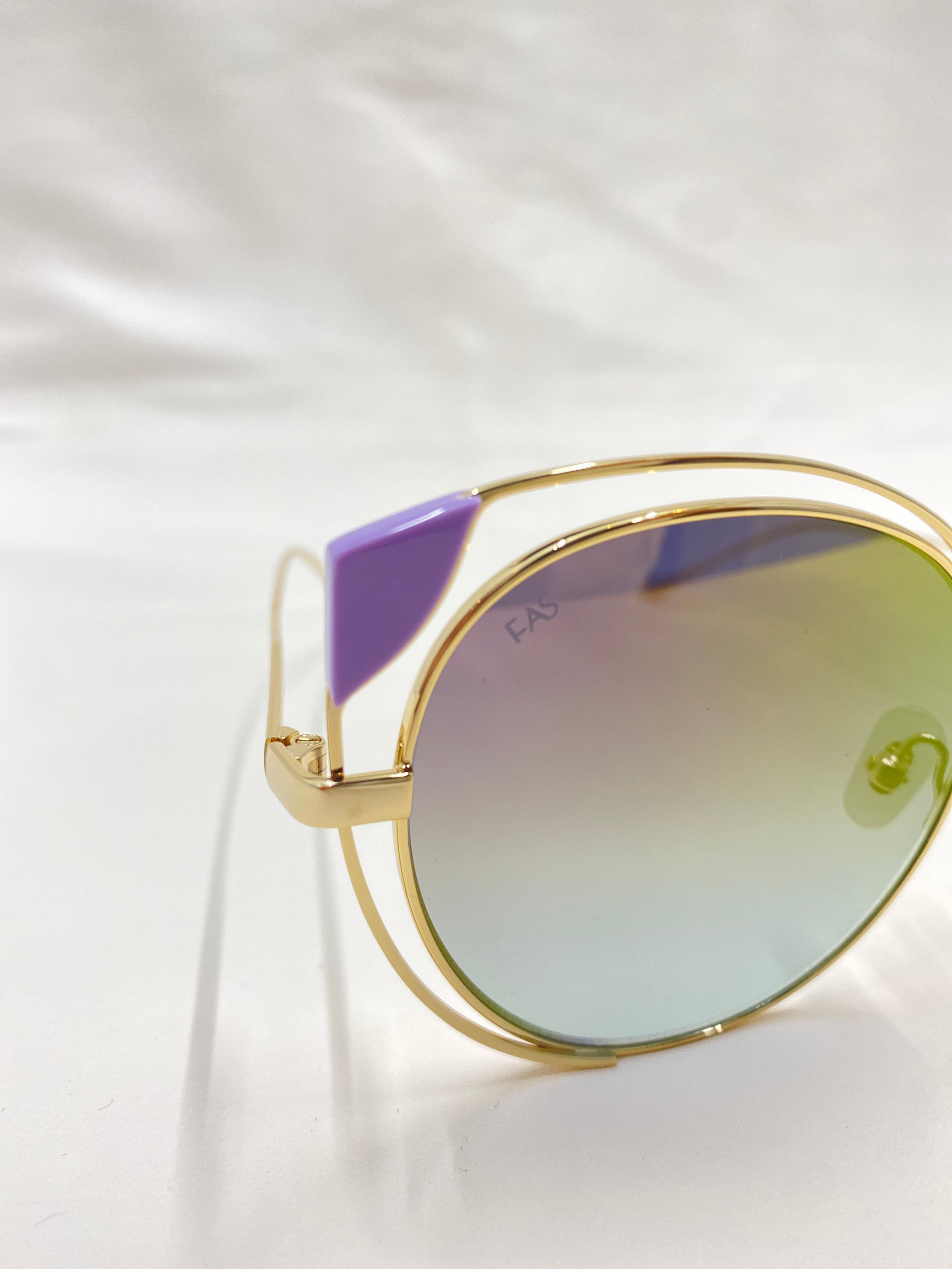 Close-up of the Skylar sunglasses by For Art's Sake® showcasing a chic cat-eye silhouette, gold-colored frames, and gradient lenses that transition from purple at the top to green at the bottom. These UVA UVB-protected sunglasses feature a purple accent on the top corner of the lens and are set against a white background.