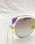 Close-up of the Skylar sunglasses by For Art's Sake® showcasing a chic cat-eye silhouette, gold-colored frames, and gradient lenses that transition from purple at the top to green at the bottom. These UVA UVB-protected sunglasses feature a purple accent on the top corner of the lens and are set against a white background.