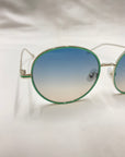 Close-up of Lemon sunglasses by For Art's Sake® featuring fashionable round gradient lenses transitioning from blue at the top to light pink at the bottom. The thin, gold-colored metal frame is adorned with hand-painted enamel detailing around the lenses and includes jadestone nose pads. The background is white.