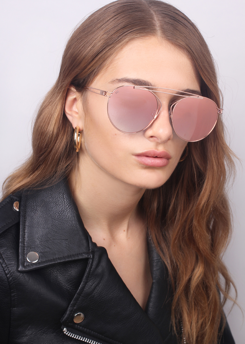 A woman with long, wavy light brown hair is wearing a For Art&#39;s Sake® T-shirt and rose-tinted aviator sunglasses with stainless steel frames. She has a black leather jacket on and is looking slightly past the camera.