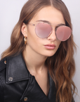 A woman with long, wavy light brown hair is wearing a For Art's Sake® T-shirt and rose-tinted aviator sunglasses with stainless steel frames. She has a black leather jacket on and is looking slightly past the camera.