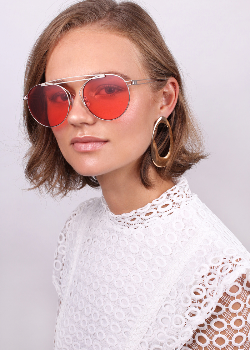 A person with short, light brown hair is adorned with large, gold hoop earrings and red-tinted aviator sunglasses featuring stainless steel frames. They are wearing a white lace top. The background is plain and light gray. The person gazes towards the camera with a calm expression, capturing the essence of For Art&#39;s Sake®.