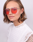 A person with short, light brown hair is adorned with large, gold hoop earrings and red-tinted aviator sunglasses featuring stainless steel frames. They are wearing a white lace top. The background is plain and light gray. The person gazes towards the camera with a calm expression, capturing the essence of For Art's Sake®.