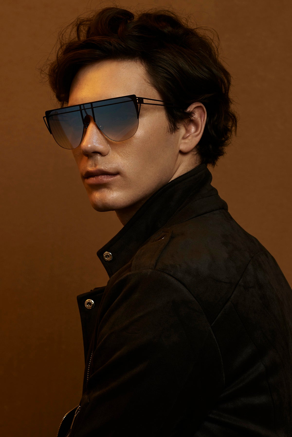 A young person with dark, wavy hair is wearing large, stylish For Art&#39;s Sake® Alien sunglasses with handmade gold-plated frames and a dark, fashionable jacket. They are posing against a plain brown background, looking slightly to the side with a calm expression.