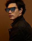 A young person with dark, wavy hair is wearing large, stylish For Art's Sake® Alien sunglasses with handmade gold-plated frames and a dark, fashionable jacket. They are posing against a plain brown background, looking slightly to the side with a calm expression.
