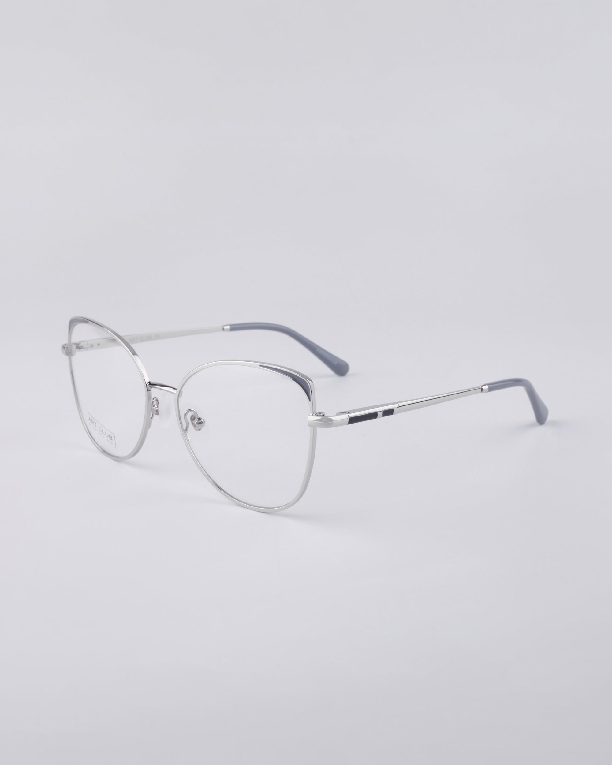 For Art's Sake® presents the Ada, a pair of minimalist silver metal eyeglasses featuring thin round frames and clear lenses. The temples, reinforced with a stainless steel core, are elegantly accented with a subtle dark hue at the tips, complementing the light gray surface.