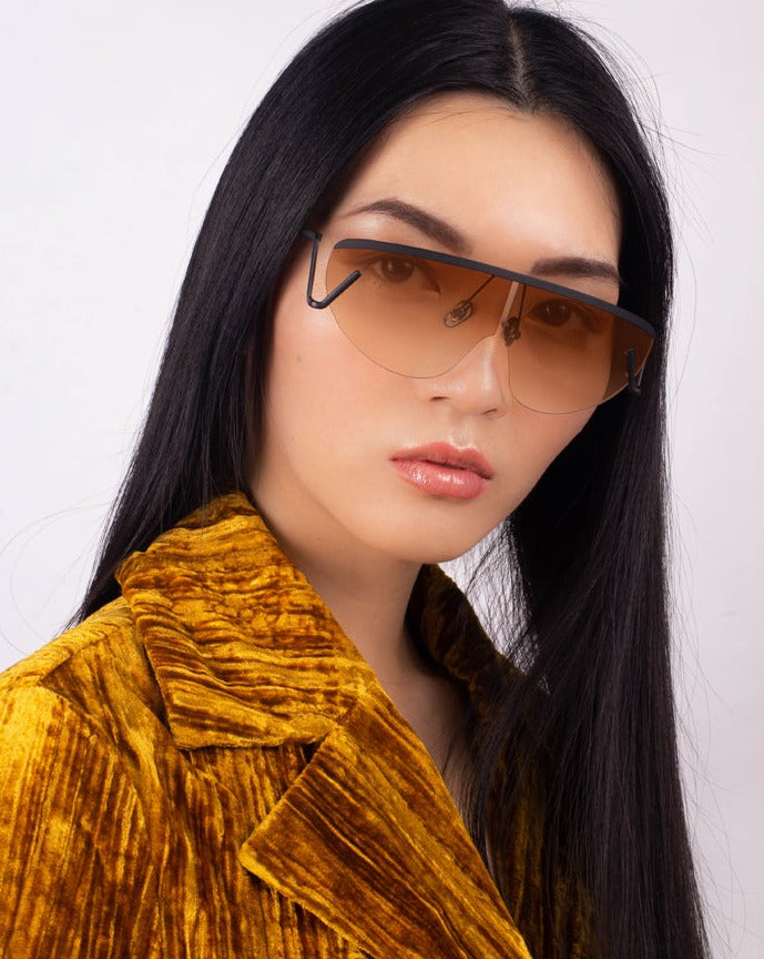 An individual with long, straight black hair is wearing For Art's Sake® Air sunglasses, featuring large, rectangular brown-tinted lenses. They are also dressed in a textured mustard yellow jacket and posing against a plain white background with a neutral expression.