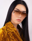 An individual with long, straight black hair is wearing For Art's Sake® Air sunglasses, featuring large, rectangular brown-tinted lenses. They are also dressed in a textured mustard yellow jacket and posing against a plain white background with a neutral expression.