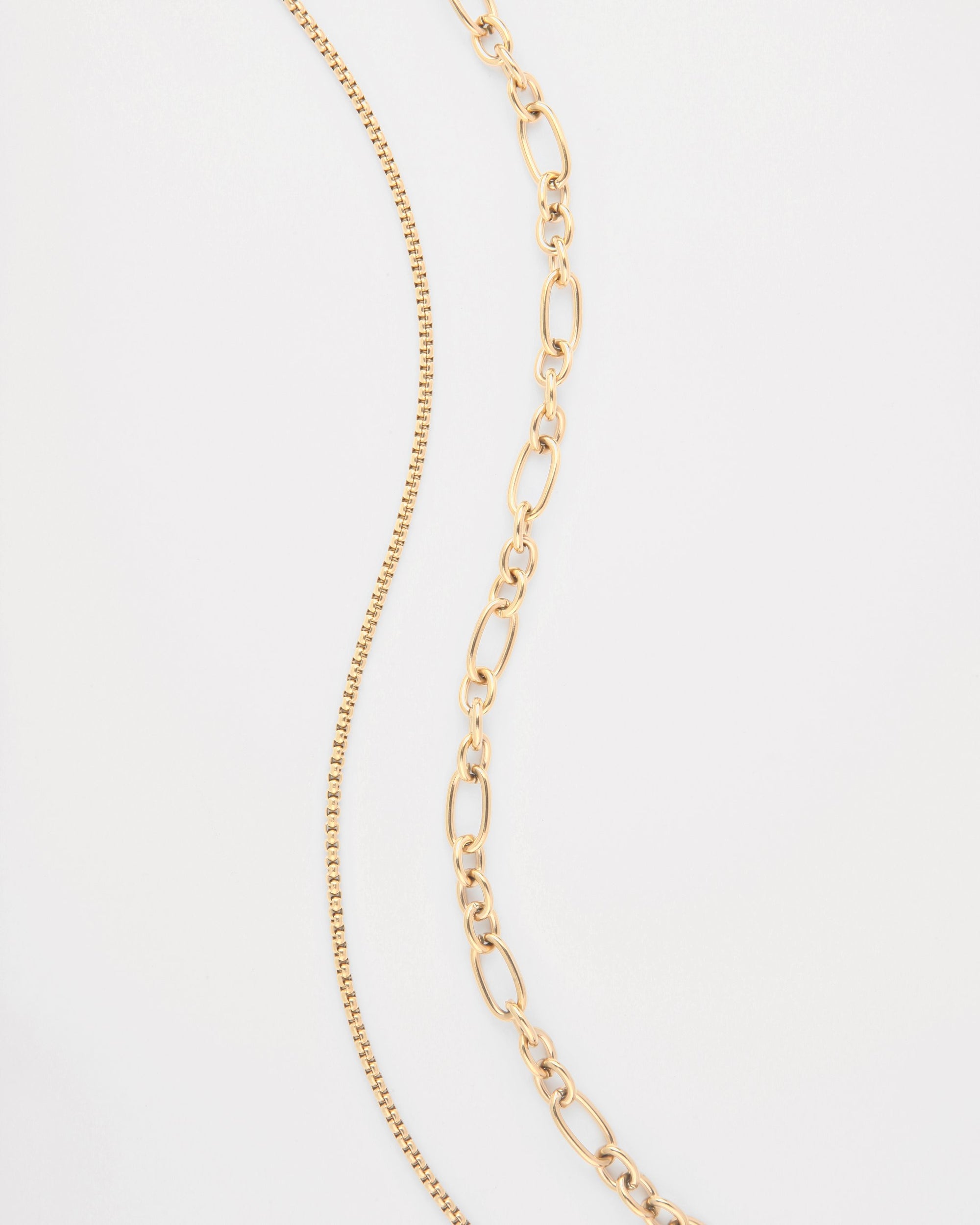 Two Alba Glasses Chains by For Art's Sake® are placed against a white background. The chain on the left, an 18K gold-plated piece, features a delicate, thin design with evenly spaced links. The chain on the right is composed of larger, oval links arranged in a slightly tapered pattern.