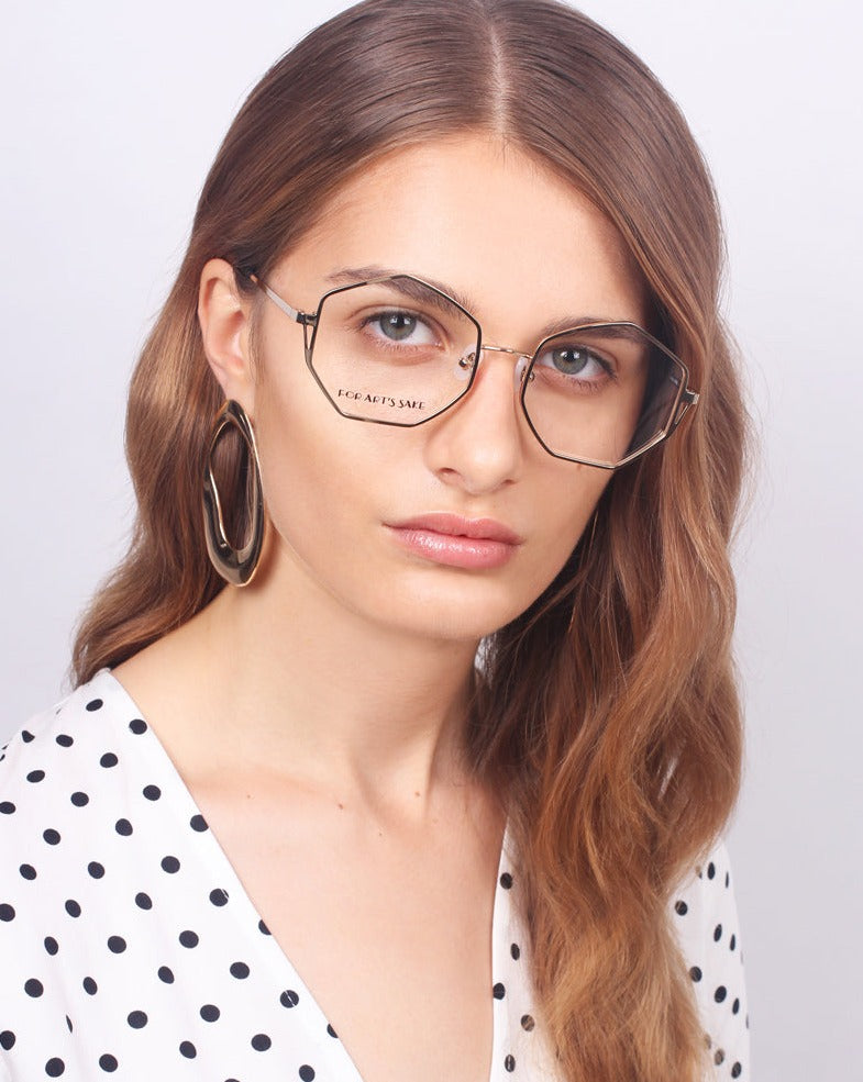 A woman with brown hair and a neutral expression is wearing geometric eyeglasses with heptagon-shaped lenses called Antidote by For Art's Sake® and large hoop earrings. She is dressed in a white blouse with black polka dots, standing against a plain background.