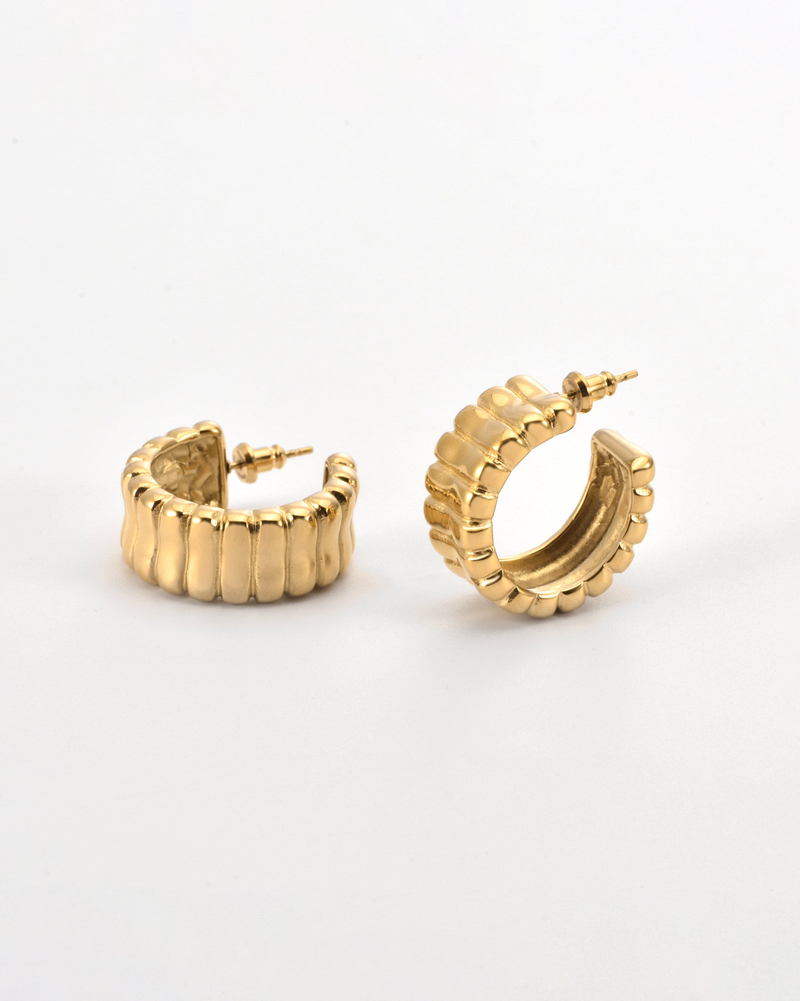 Close-up of a pair of For Art&#39;s Sake® Arch Earrings Gold with a ribbed texture. One earring is laying flat, while the other stands upright, showcasing their semi-circular design with a modern edge. The background is a plain, light surface.