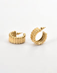 Close-up of a pair of For Art's Sake® Arch Earrings Gold with a ribbed texture. One earring is laying flat, while the other stands upright, showcasing their semi-circular design with a modern edge. The background is a plain, light surface.