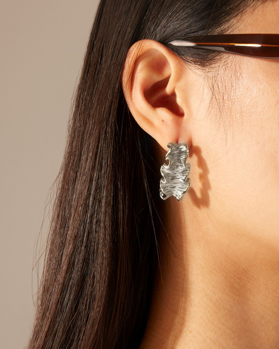 Close-up of a person with long dark hair, wearing large, handcrafted For Art's Sake® Athena Earrings Silver shaped like abstract leaves. The person is seen from the side, highlighting the earring. Their eyeglasses and part of their ear are also visible. The background is neutral.