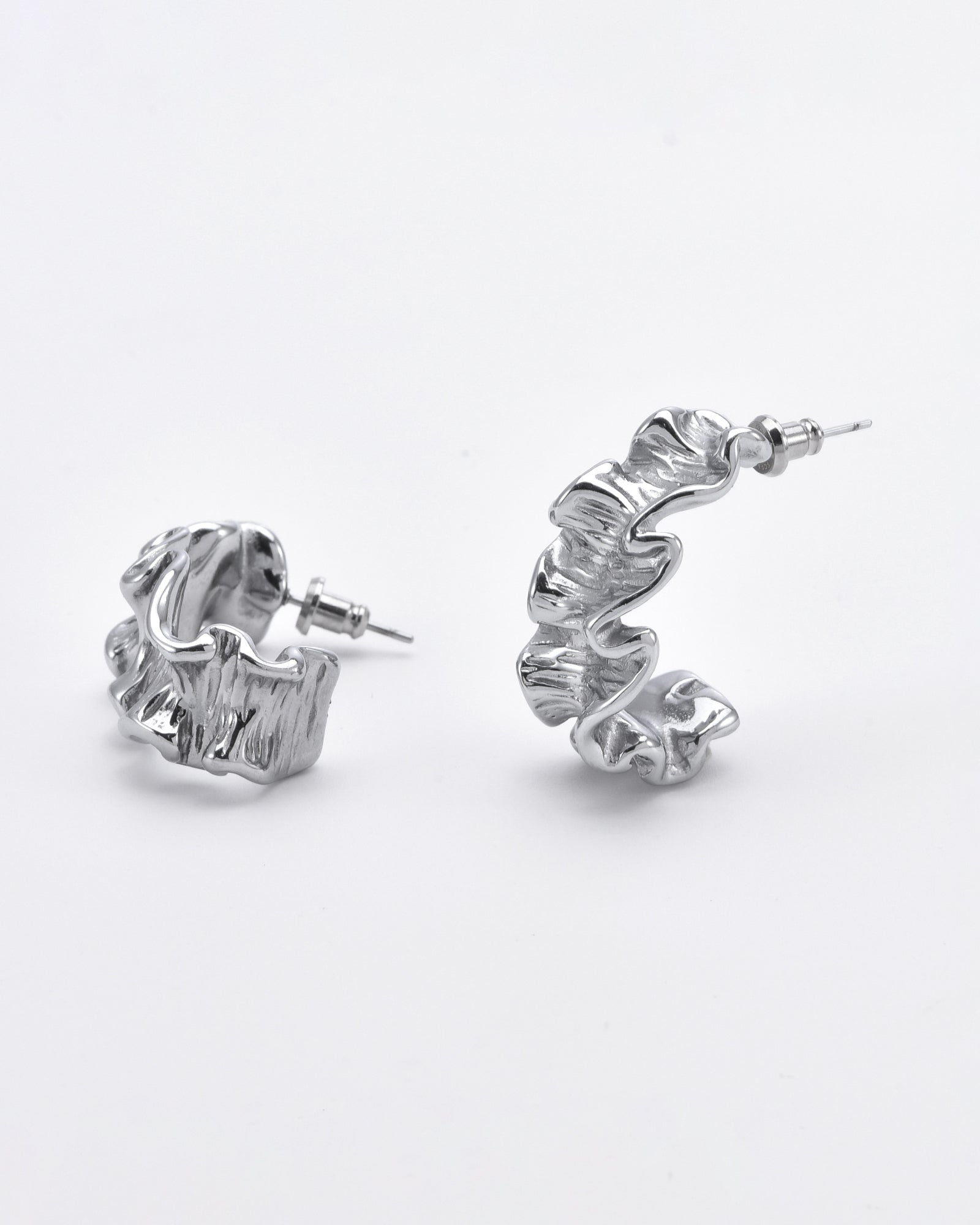A pair of handcrafted silver hoop earrings with a ruffled, textured design. One earring is standing upright, while the other lies flat, showcasing their intricate, wavy patterns on a white background. These Athena Earrings Silver by For Art's Sake® exude elegance and artistry in every detail.
