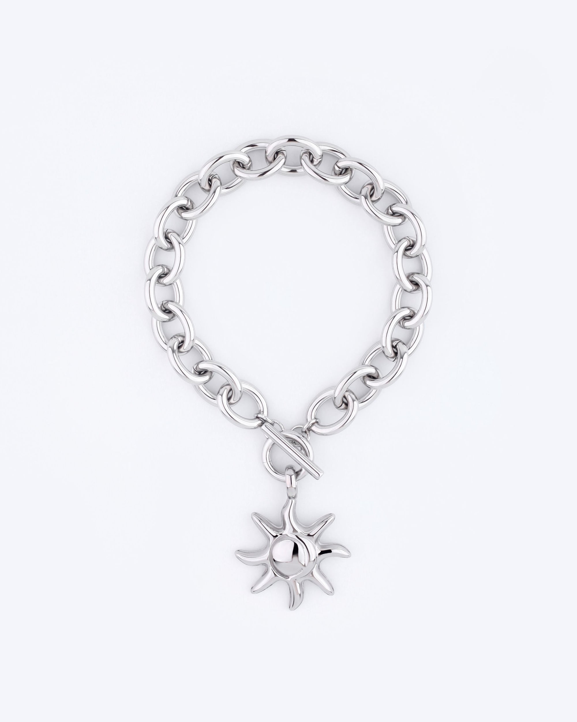 The Crescent Moon Bracelet by For Art's Sake® is a silver chain bracelet with a circular toggle clasp, featuring a sun-shaped charm that hangs elegantly. The charm's rays and polished surface exude an elegant charisma. With a 20cm length, this exquisite piece is beautifully set against a simple, light grey background.