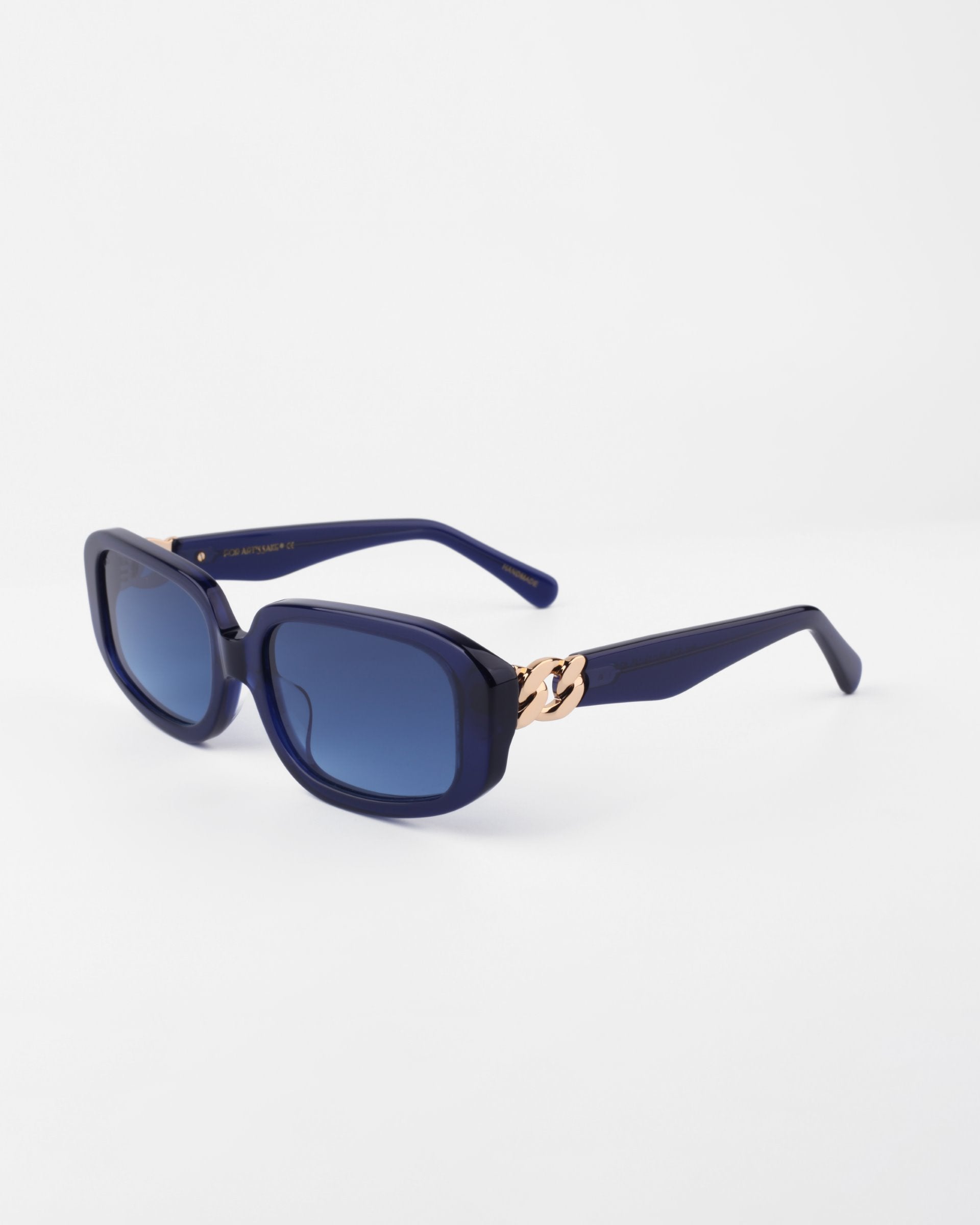 A pair of stylish navy blue For Art&#39;s Sake® Bolt sunglasses with square lenses and dark blue tinted, shatter-resistant nylon glasses. The thick handmade acetate frame features gold-plated temple detailing near the hinges. The background is plain white.