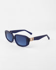 A pair of stylish navy blue For Art's Sake® Bolt sunglasses with square lenses and dark blue tinted, shatter-resistant nylon glasses. The thick handmade acetate frame features gold-plated temple detailing near the hinges. The background is plain white.