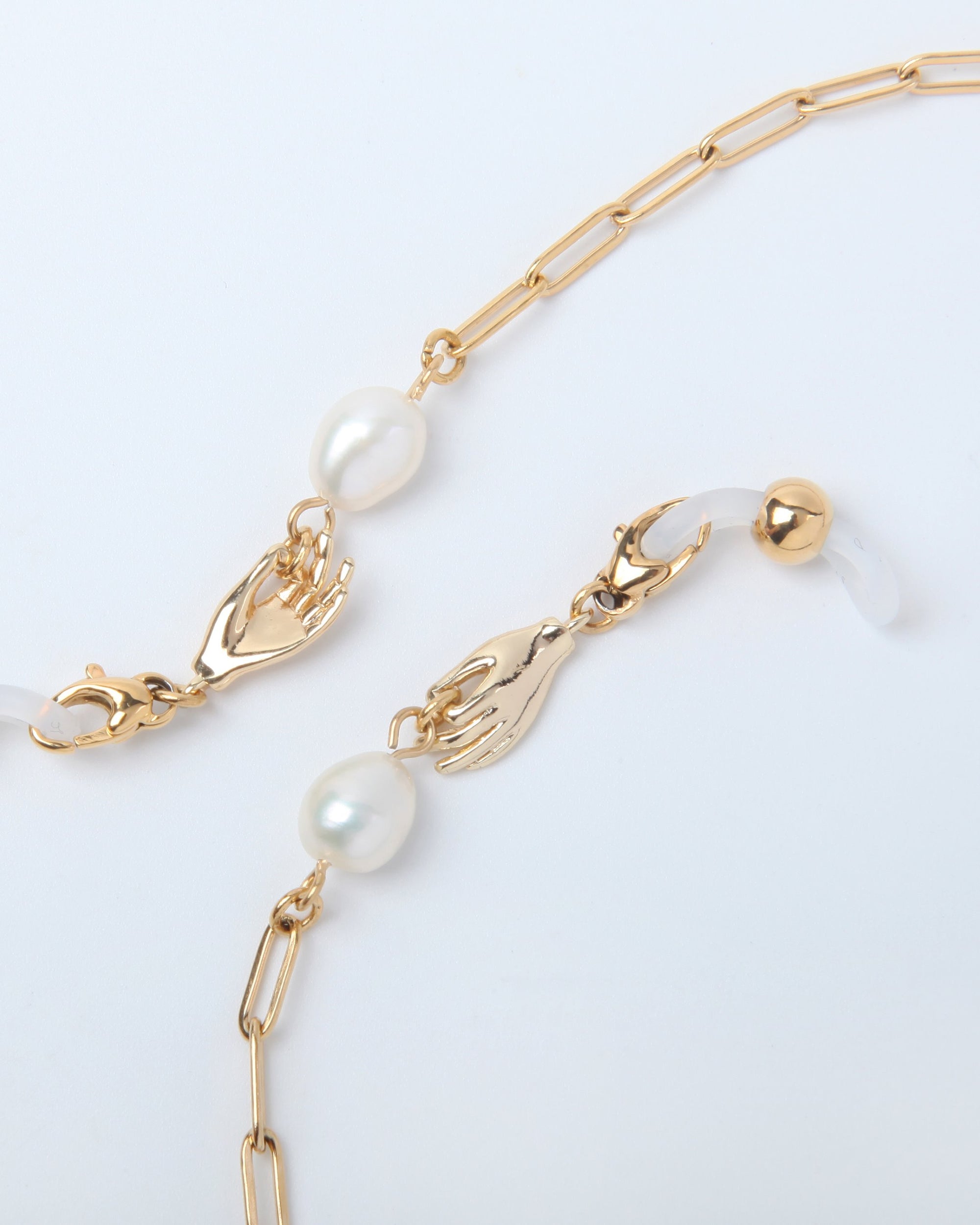 The For Art's Sake® Buddha Glasses Chain is a delicate 18-karat gold-plated chain necklace with two small hands clasping parts of the chain, each holding a white freshwater pearl. The chain features elongated links, and the hands serve as unique decorative elements. One end of the chain has a round gold bead.