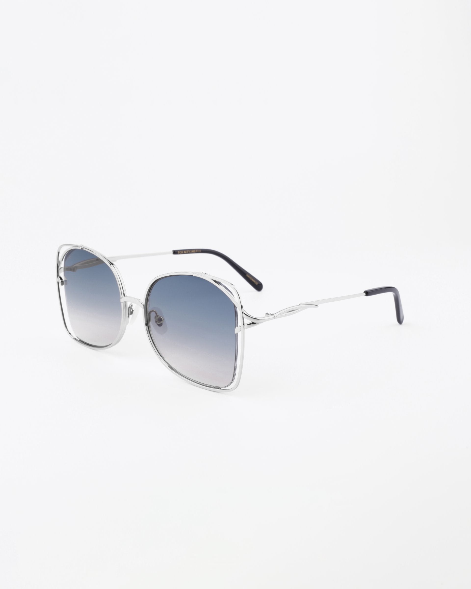 A pair of stylish handmade sunglasses, Carousel by For Art's Sake®, with shatter-resistant, large rounded square blue gradient lenses and thin temples that extend into black tips. The silver metal frame adds a sophisticated touch against the plain white background.