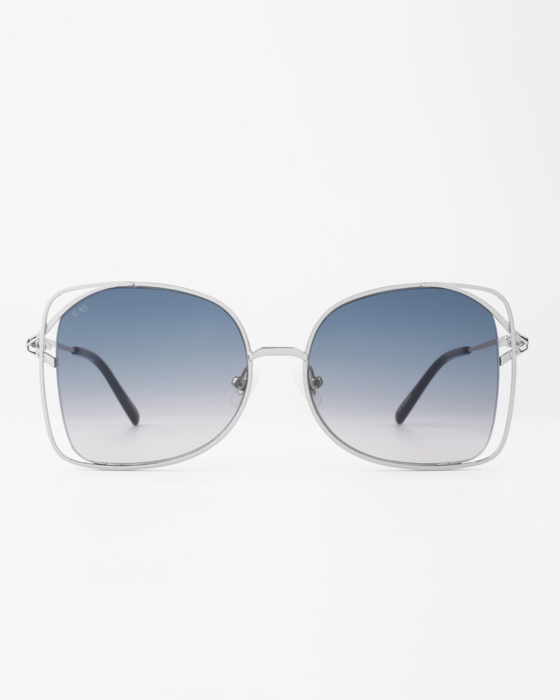 A pair of oversized, handmade Carousel sunglasses from For Art&#39;s Sake® with rounded square frames made of thin silver metal. The gradient lenses transition from dark blue at the top to clear at the bottom, providing UVA &amp; UVB protection. The sunglasses feature thin arms with black tips. The background is plain white.