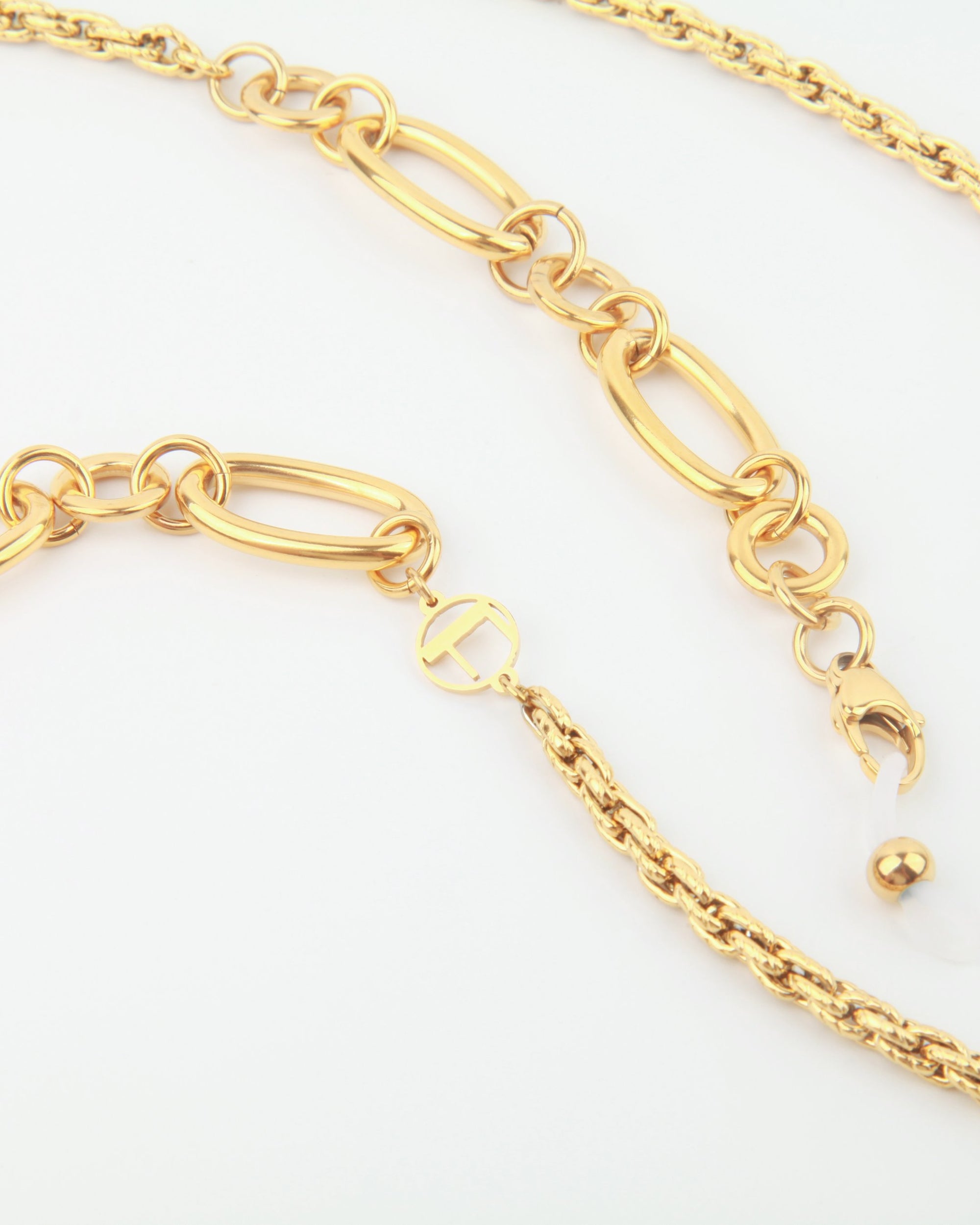 A close-up image of a gold-plated stainless steel Milan Glasses Chain by For Art's Sake® with a combination of large oval and smaller round links. The chain features a circular clasp with a decorative "T" charm. The ornate chain has a glossy finish and rests on a white background.