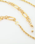 A close-up image of a gold-plated stainless steel Milan Glasses Chain by For Art's Sake® with a combination of large oval and smaller round links. The chain features a circular clasp with a decorative "T" charm. The ornate chain has a glossy finish and rests on a white background.