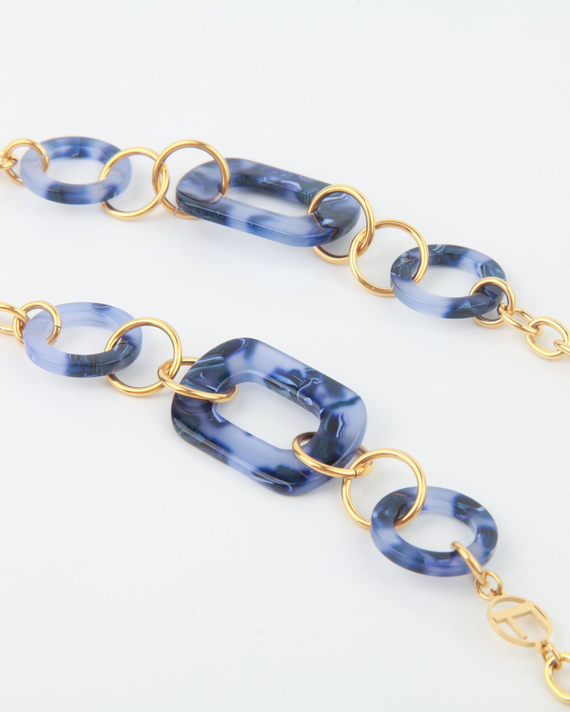 A close-up of a fashionable bracelet featuring 18-karat gold-plated links and marbled blue resin rings. The design alternates between circular and rectangular resin pieces, connected by bright gold loops, creating a modern For Art&#39;s Sake® NYC Glasses Chain-style accessory.