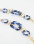 A close-up of a fashionable bracelet featuring 18-karat gold-plated links and marbled blue resin rings. The design alternates between circular and rectangular resin pieces, connected by bright gold loops, creating a modern For Art's Sake® NYC Glasses Chain-style accessory.