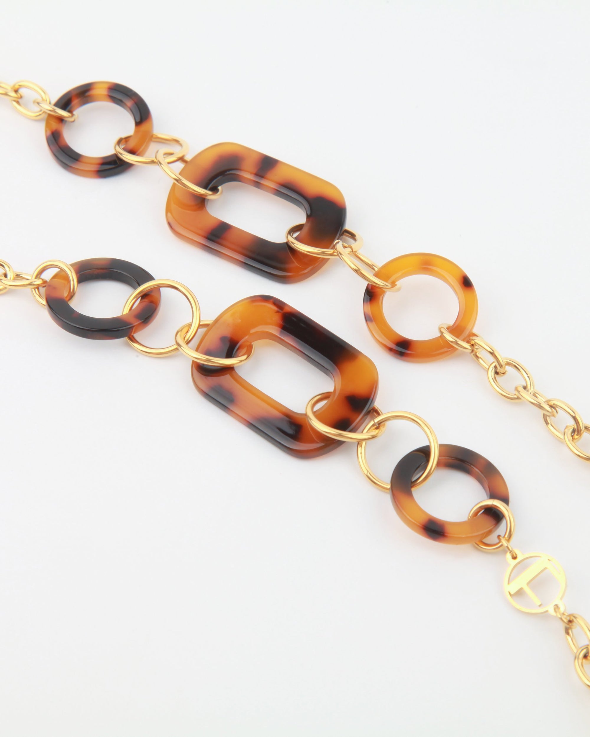 A close-up view of a stylish chain necklace featuring alternating large tortoiseshell oval links and smaller circular links, connected by 18-karat gold-plated rings. The NYC Glasses Chain by For Art&#39;s Sake® has a sophisticated, modern design with oversized acetate links and is displayed against a plain white background.