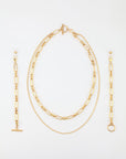 A Vienna Glasses Chain by For Art's Sake® against a white background, including a pair of dangling earrings, an 18-karat gold-plating layered necklace with both a chain and a linked design, and a bracelet featuring a similar linked pattern. The pieces are neatly arranged in a vertical orientation.
