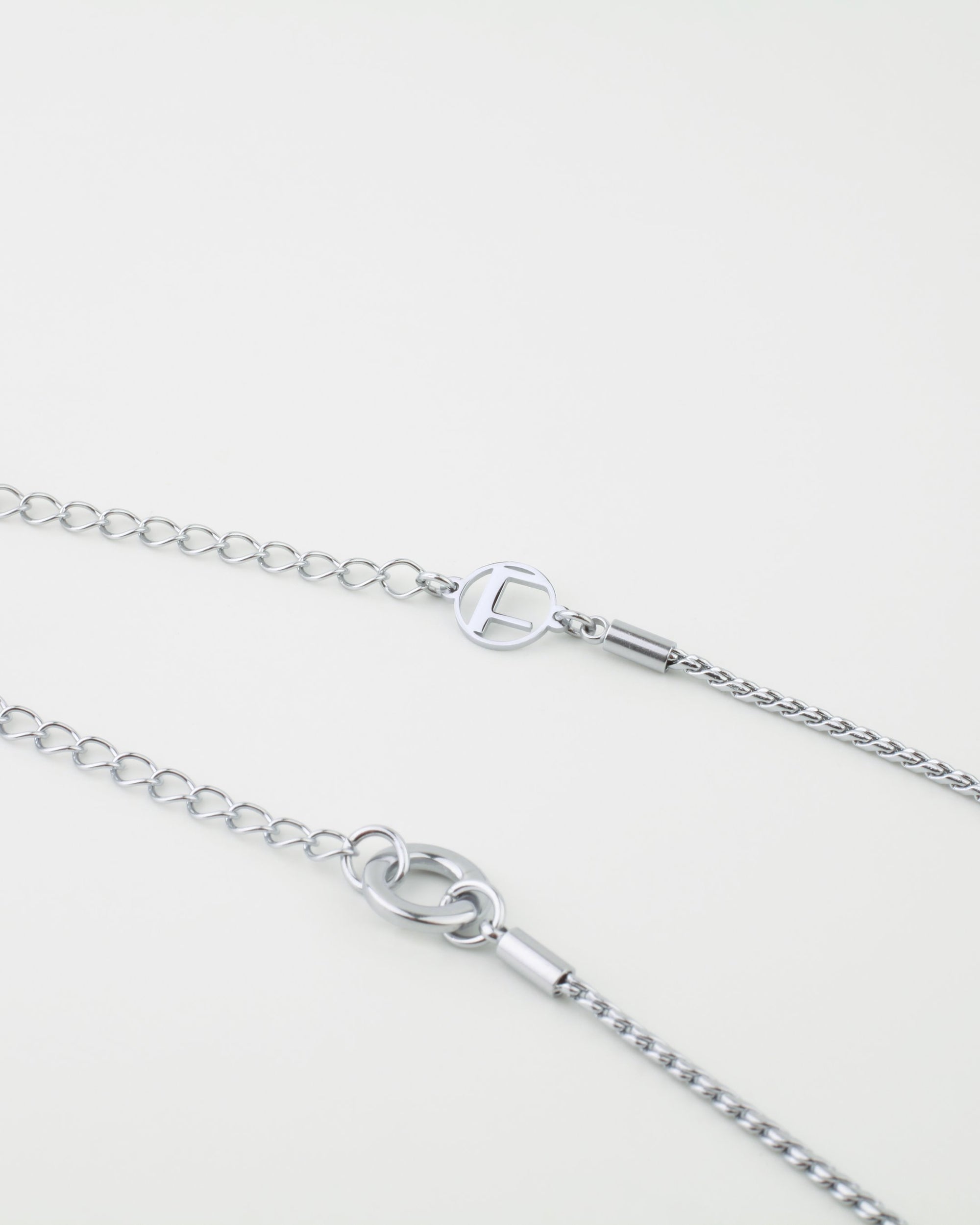 Close-up of two silver necklaces lying parallel against a light background. Each necklace has a thin wheat chain with different styled clasp mechanisms and additional chain link closures for adjustable lengths. The upper necklace features a circular clasp with an "F" inside, part of the Miami Glasses Chain by For Art's Sake®.