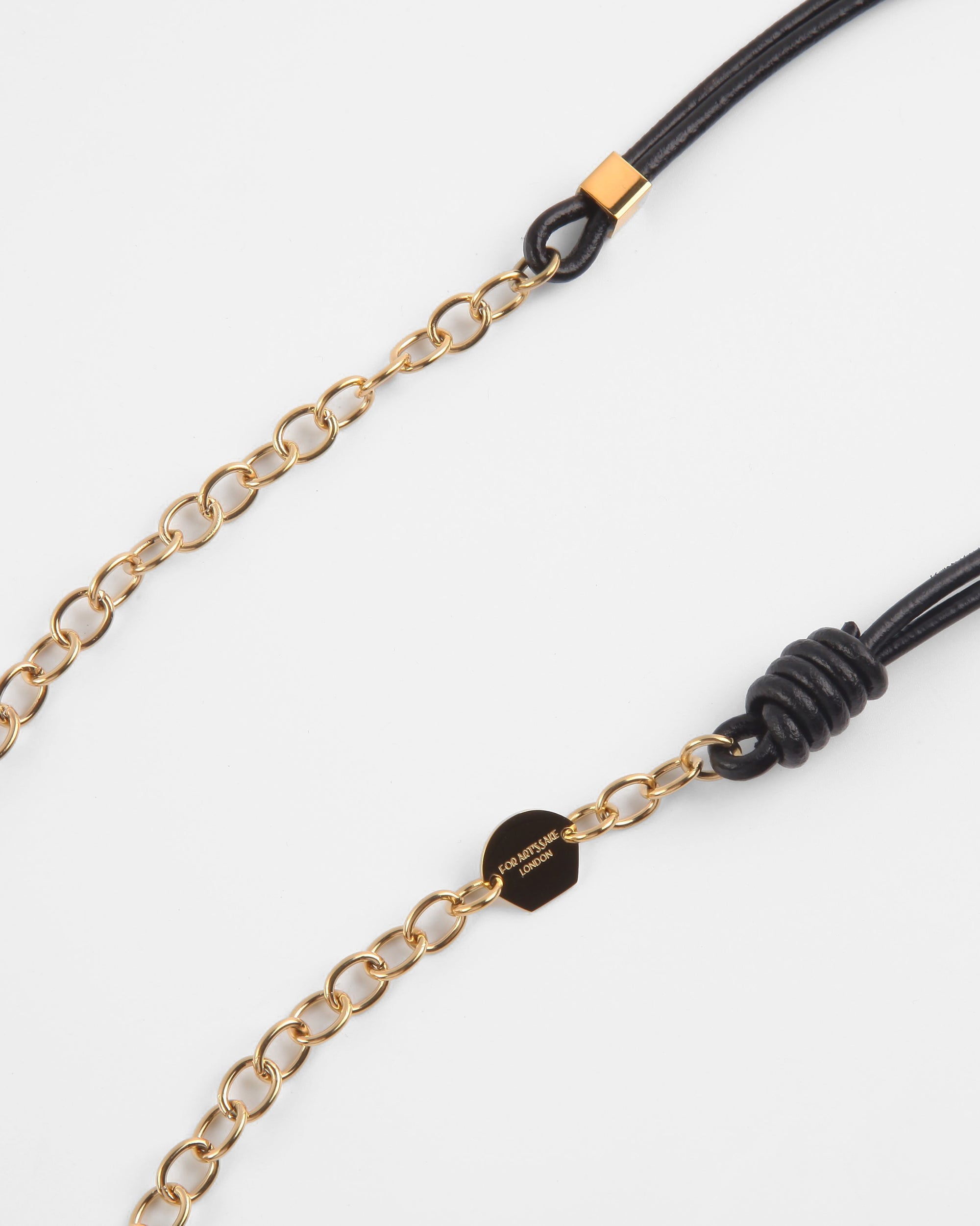 A close-up shot of a Soho Glasses Chain with 18-Karat gold links and premium leather adjustable cords by For Art's Sake®. The black leather cords feature gold-tone hardware details at each end. One end has a small gold tag with engraved text, while the other includes a knotted leather loop. The background is plain white.