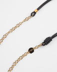 A close-up shot of a Soho Glasses Chain with 18-Karat gold links and premium leather adjustable cords by For Art's Sake®. The black leather cords feature gold-tone hardware details at each end. One end has a small gold tag with engraved text, while the other includes a knotted leather loop. The background is plain white.