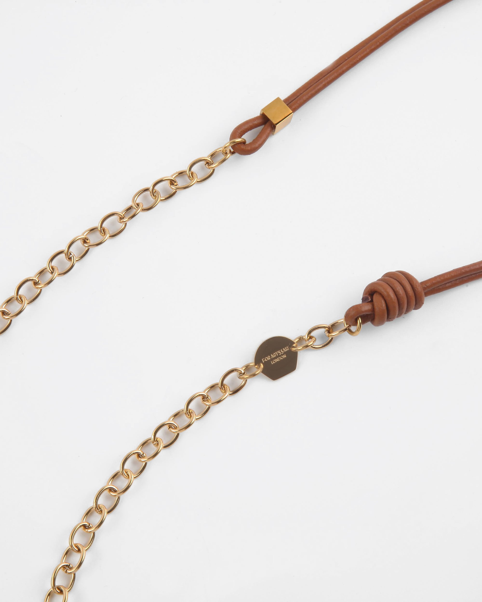 Close-up of a brown premium leather strap attached to 18-karat gold links. The strap is looped and secured with a knot on one end and a gold buckle on the other. A gold tag is attached to the chain with engraved text. The background is plain white, showcasing this elegant Soho Glasses Chain by For Art's Sake®.