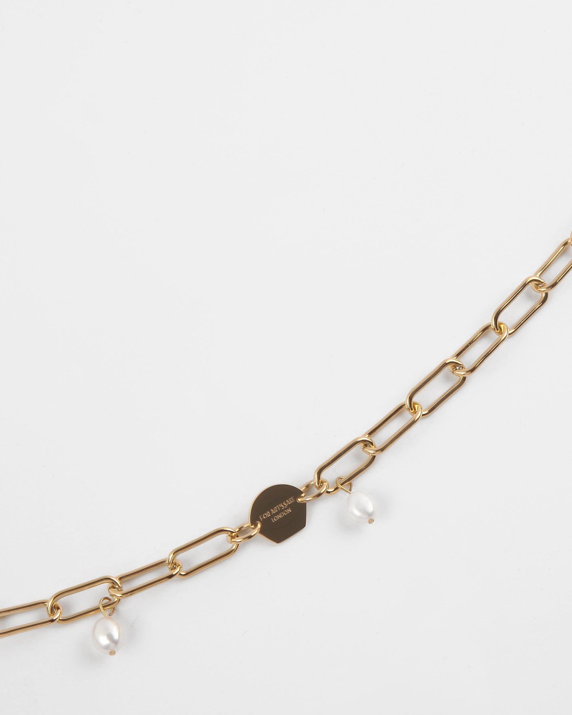 A delicate 18K gold-plated Marylebone Glasses Chain featuring elongated links, adorned with small freshwater pearl pendants. A For Art's Sake® charm is attached to the chain, displaying text that appears to be a brand name or logo. The background is a plain white surface.