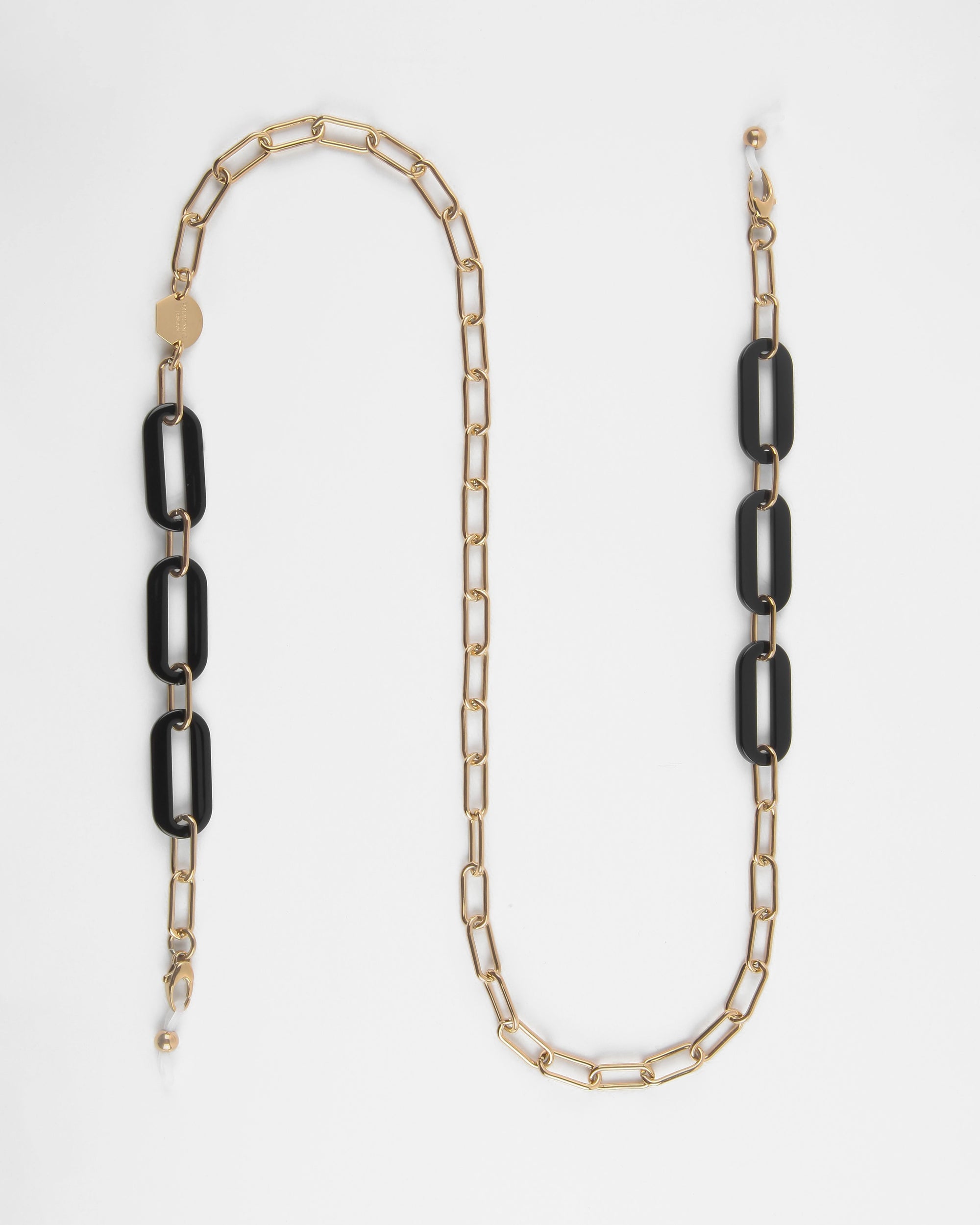 A Kensington Glasses Chain featuring an alternating pattern of rectangular black links and Italian acetate on one side. The For Art&#39;s Sake® 18K gold-plated chain is laid out in a relaxed, slightly curved manner against a white background.