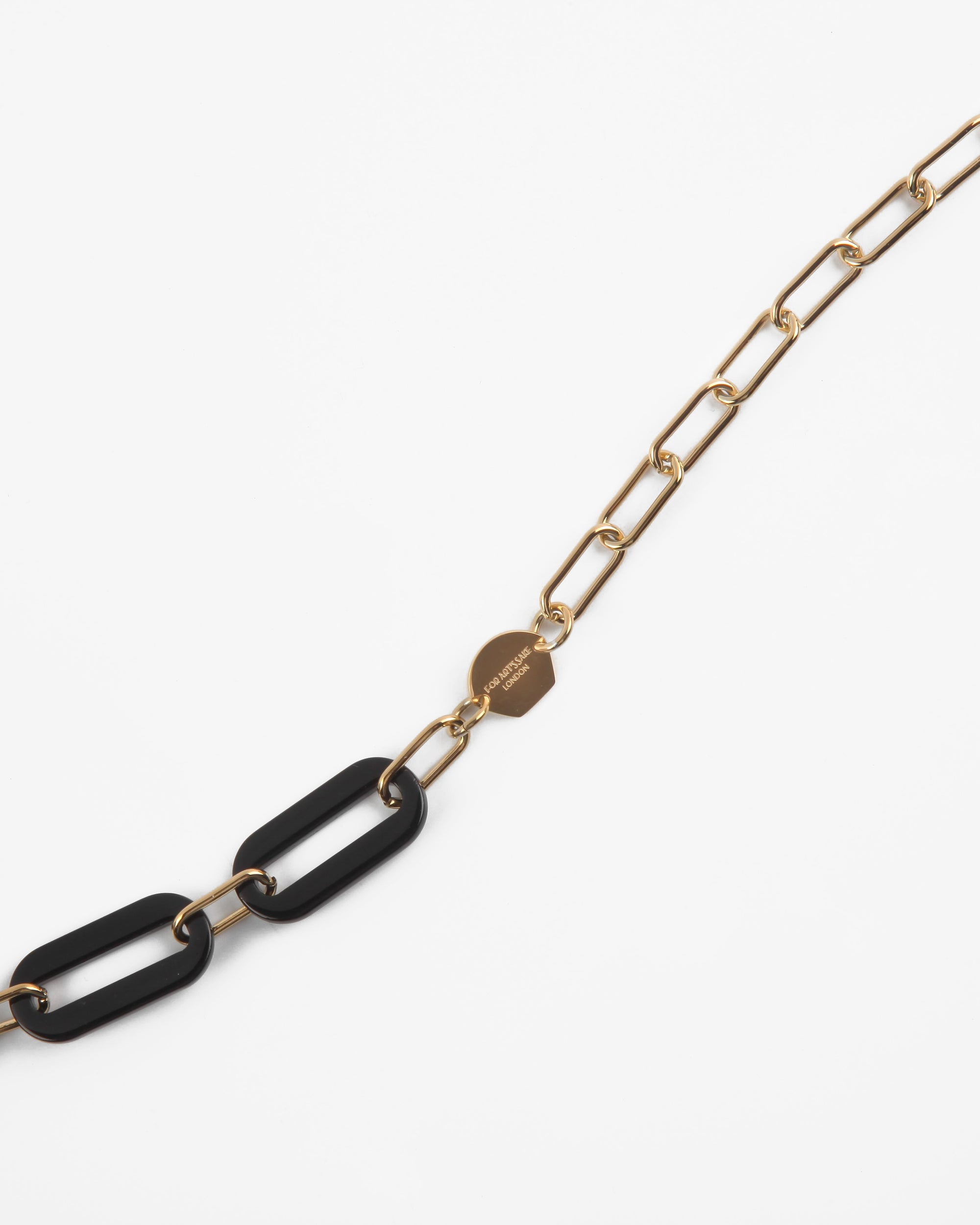 A chic, minimalist necklace featuring a chain with elongated, 18K gold-plated links. In the center are two large, black rectangular links made from Italian acetate. A small, gold circular pendant with an engraved design is attached to the chain near the black links, adding a stylish touch. This is the Kensington Glasses Chain by For Art&#39;s Sake®.